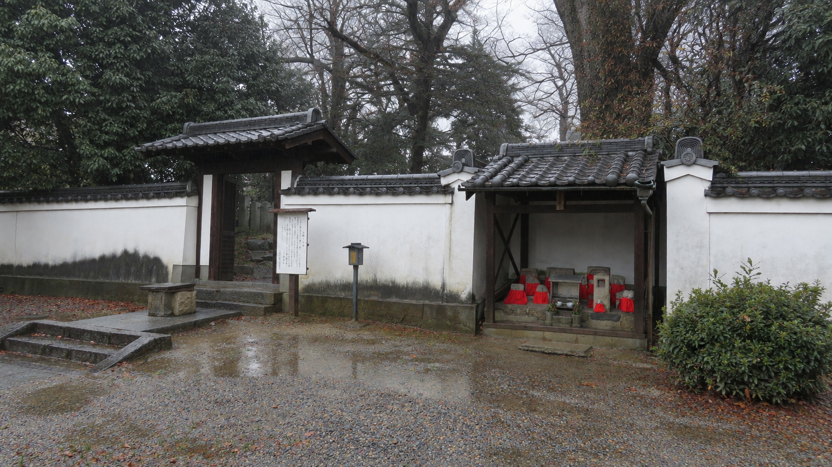 塀に囲まれた入口の門。右手には赤い前掛けの地蔵が並ぶ祠があります